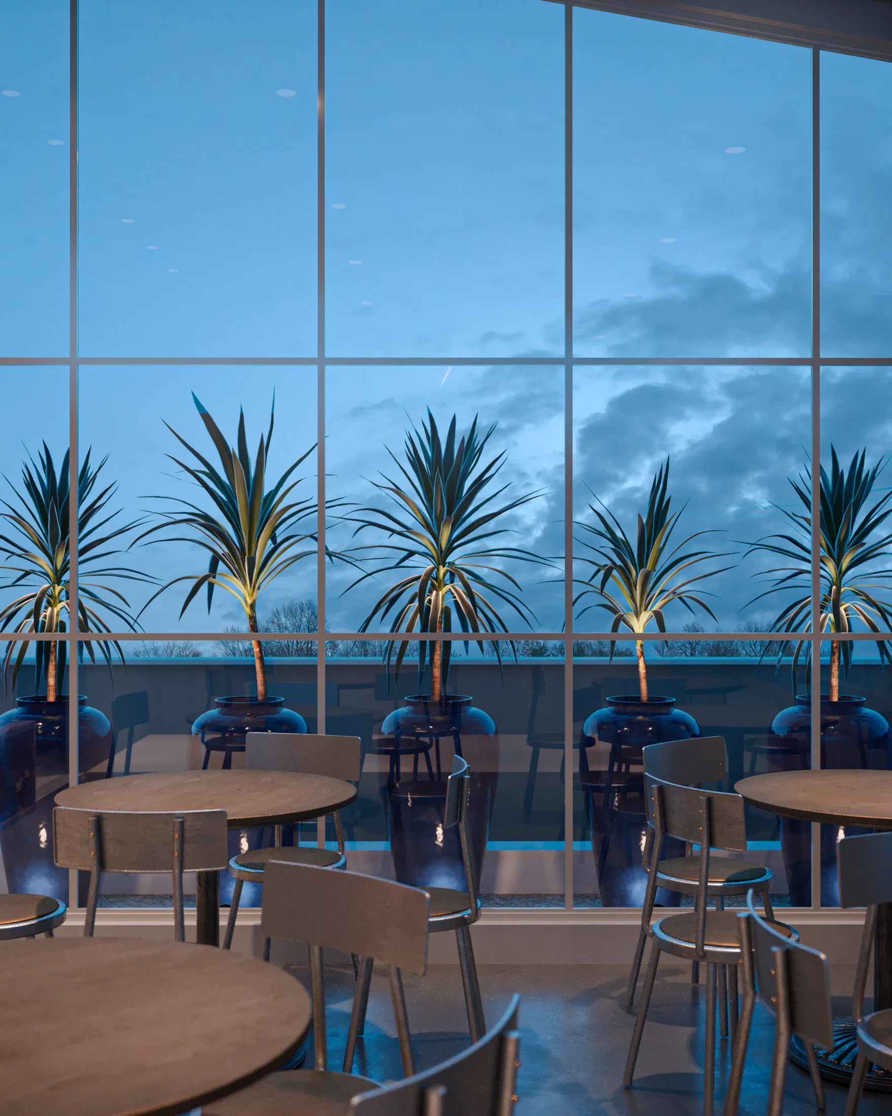 Salão interno ao entardecer, com mesas redondas de madeira e vista através de fachada envidraçada para vasos azuis com dracenas em terraço externo